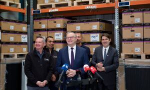 Group of men in a warehouse press conference, with stacked Fox C07 battery boxes behind them. Group of men in a warehouse press conference, with stacked Fox C07 battery boxes behind them.
