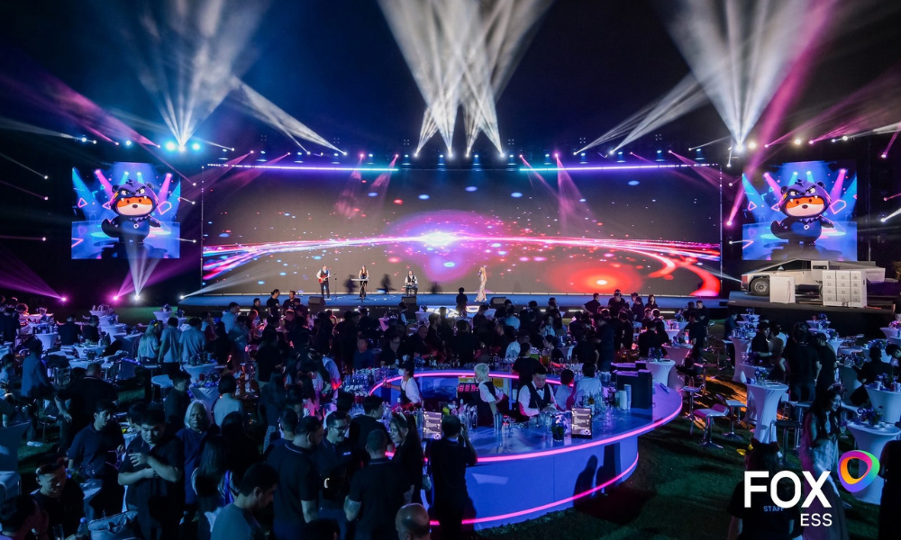 Wide shot of a large night-time event with performers on a bright stage, colorful laser lighting, and a seated audience around circular bars; FOX ESS branding visible. Wide shot of a large night-time event with performers on a bright stage, colorful laser lighting, and a seated audience around circular bars; FOX ESS branding visible.