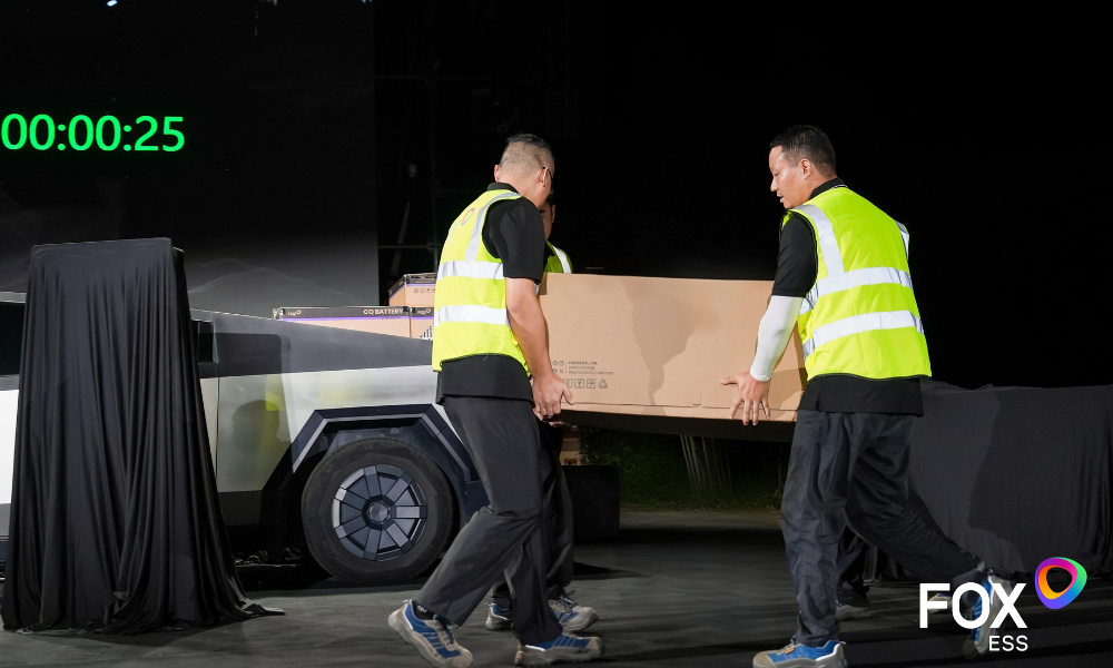 Two workers in high-visibility vests carry a large cardboard box onto a stage beside a futuristic vehicle as a FOX ESS presentation unfolds in the background. Two workers in high-visibility vests carry a large cardboard box onto a stage beside a futuristic vehicle as a FOX ESS presentation unfolds in the background.