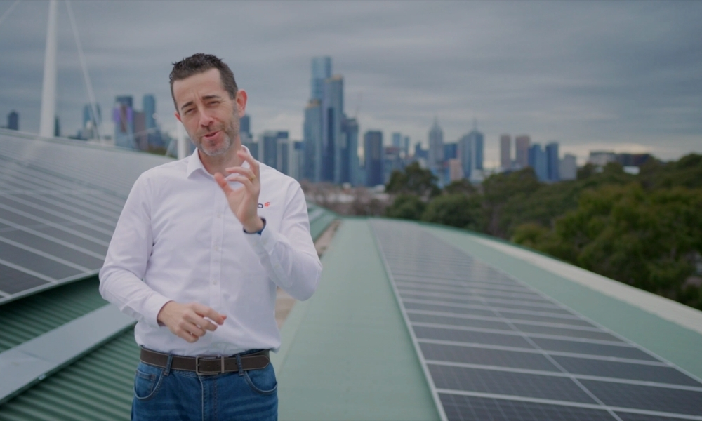 Man in a white shirt and jeans speaks on a rooftop, gesturing, with solar panels and a city skyline in the background. Man in a white shirt and jeans speaks on a rooftop, gesturing, with solar panels and a city skyline in the background.
