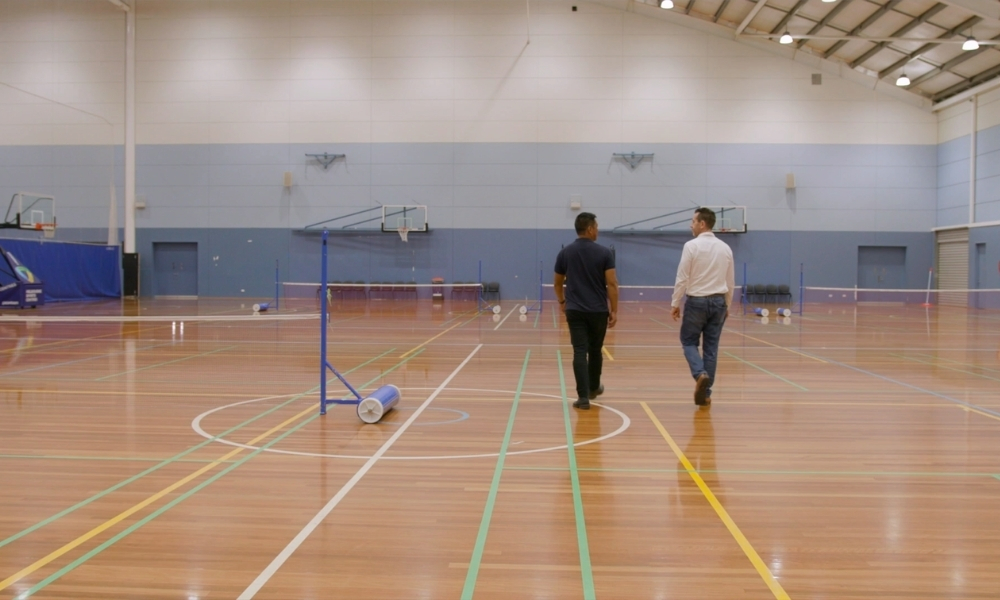 Two men walk away in a large indoor gym with badminton nets and equipment on a polished wooden floor. Two men walk away in a large indoor gym with badminton nets and equipment on a polished wooden floor.
