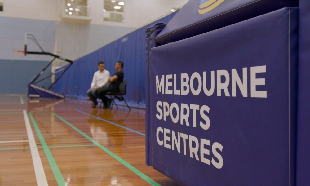 Two men sit and chat inside a gym beside a large blue padded barrier labeled 'Melbourne Sports Centres' with a basketball hoop in the background. Two men sit and chat inside a gym beside a large blue padded barrier labeled 'Melbourne Sports Centres' with a basketball hoop in the background.