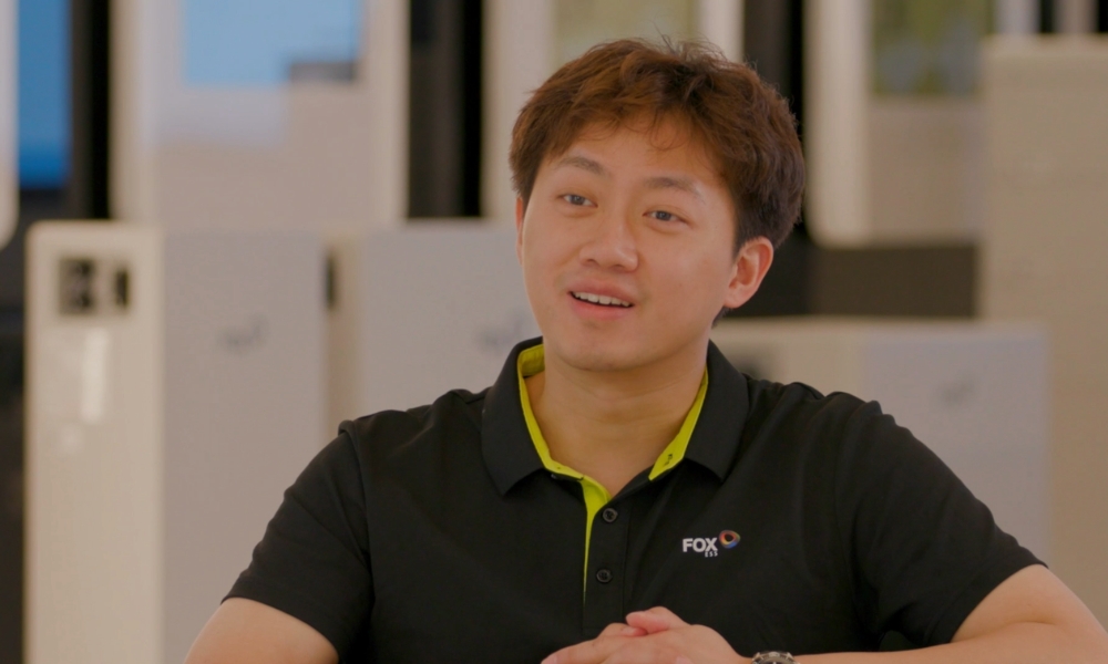 Young man in a black polo with lime-green collar sits at a desk, talking with folded hands. Young man in a black polo with lime-green collar sits at a desk, talking with folded hands.