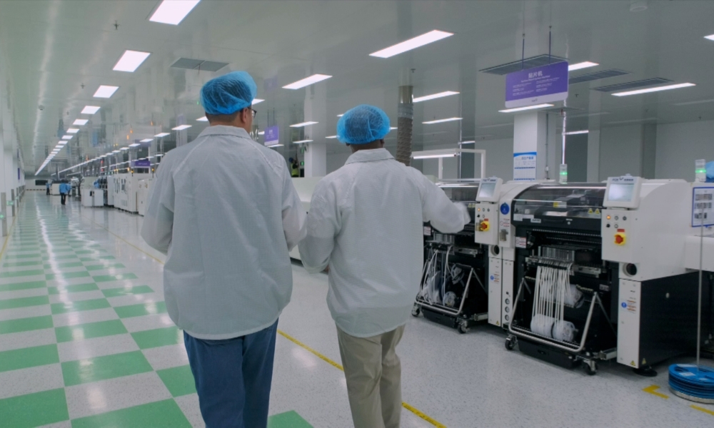 Two workers in white coats and blue hairnets walk down a cleanroom floor beside automated manufacturing machines. Two workers in white coats and blue hairnets walk down a cleanroom floor beside automated manufacturing machines.