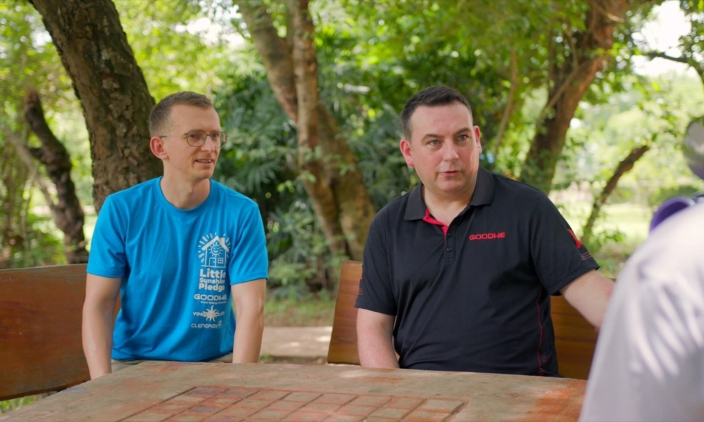 Two men sit at a wooden outdoor table in a park, one in a blue shirt with glasses, the other in a black polo, engaged in conversation. Two men sit at a wooden outdoor table in a park, one in a blue shirt with glasses, the other in a black polo, engaged in conversation.
