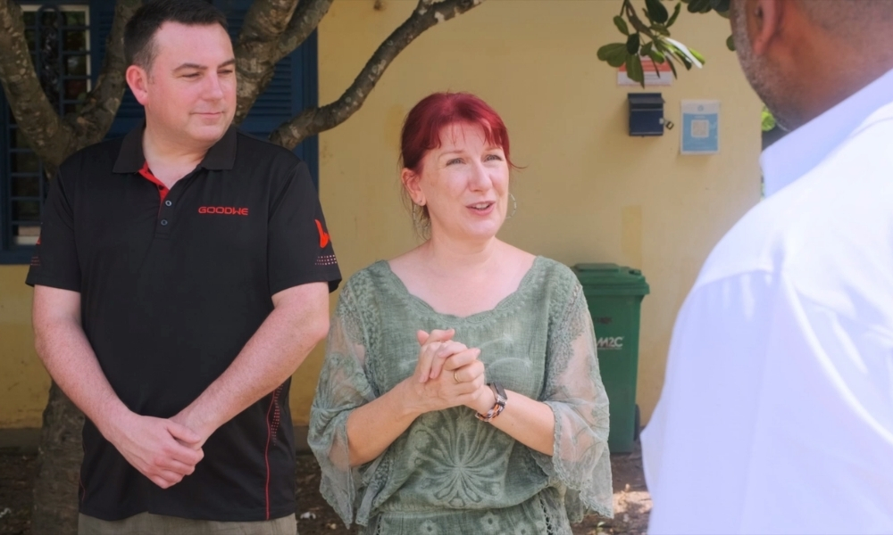 Two people converse outdoors: a man in a black polo and a red-haired woman in a green top, with a third person partially visible on the right. Two people converse outdoors: a man in a black polo and a red-haired woman in a green top, with a third person partially visible on the right.