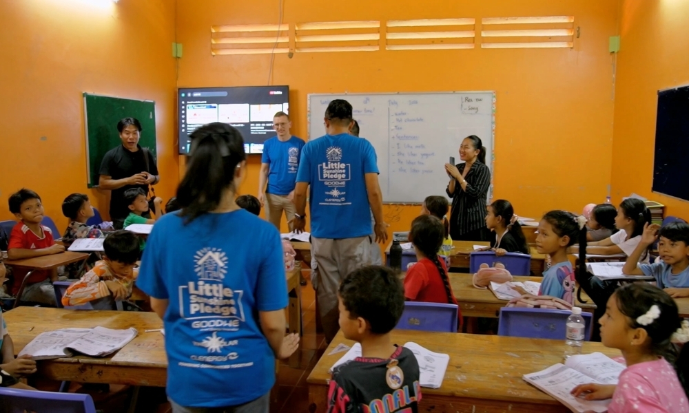 Volunteer teachers in blue shirts help a classroom of children in an orange-walled room with a whiteboard and a large screen at the front. Volunteer teachers in blue shirts help a classroom of children in an orange-walled room with a whiteboard and a large screen at the front.