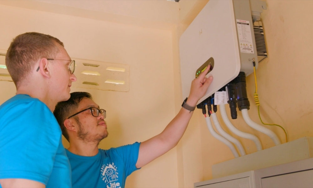 Two technicians in blue shirts install or adjust a wall-mounted electrical inverter/energy management unit, connecting multiple white cables beneath it. Two technicians in blue shirts install or adjust a wall-mounted electrical inverter/energy management unit, connecting multiple white cables beneath it.