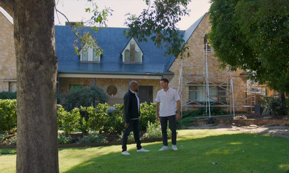 Two men stand on a green lawn in front of a stone house with a blue roof and scaffolding on the right side of the yard. Two men stand on a green lawn in front of a stone house with a blue roof and scaffolding on the right side of the yard.