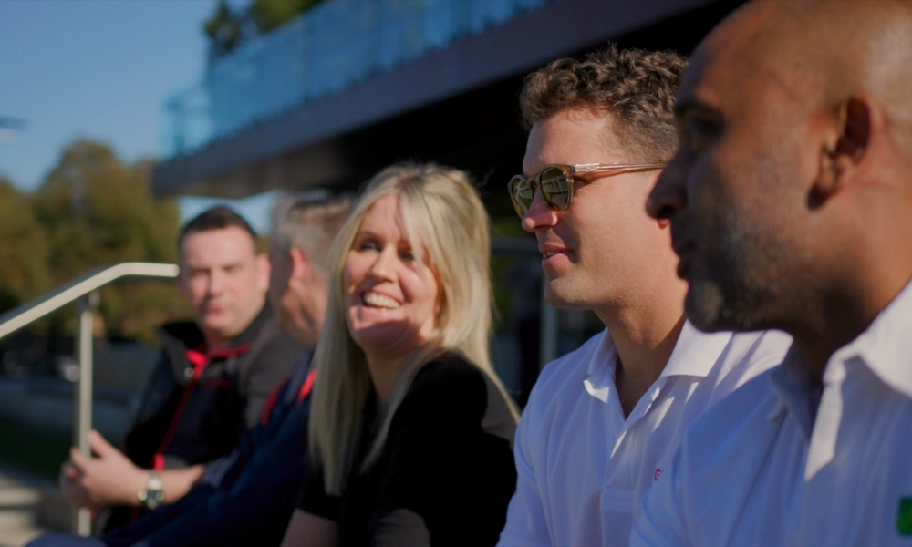Four adults sitting on a bench outdoors, smiling and chatting; focus on a man in a white shirt wearing sunglasses in the foreground. Four adults sitting on a bench outdoors, smiling and chatting; focus on a man in a white shirt wearing sunglasses in the foreground.