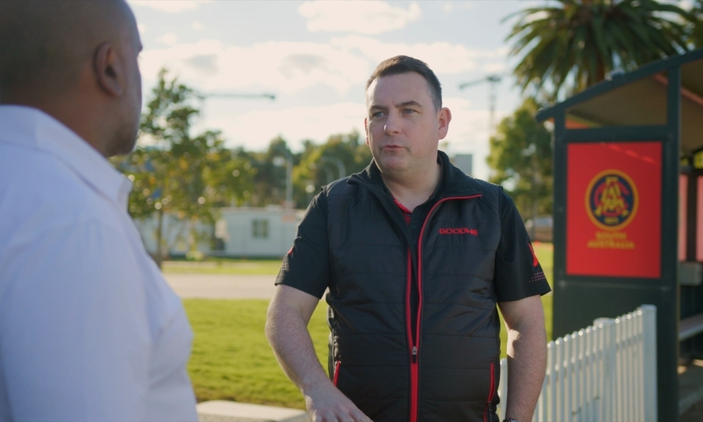 Two men talking outdoors; one facing the camera in a black vest with red zippers, the other in a white shirt. Two men talking outdoors; one facing the camera in a black vest with red zippers, the other in a white shirt.