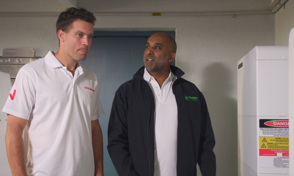 Two men in a utility room stand and talk near a large white machine with a danger label, one in a white polo and the other in a black jacket. Two men in a utility room stand and talk near a large white machine with a danger label, one in a white polo and the other in a black jacket.