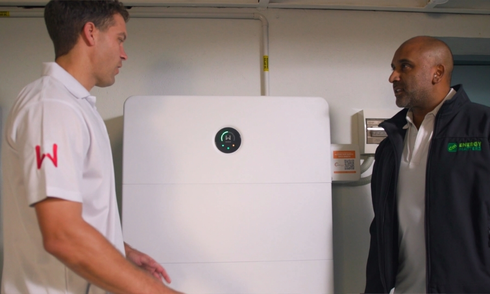 Two men in a utility room stand facing each other beside a large white industrial device with a circular display panel. Two men in a utility room stand facing each other beside a large white industrial device with a circular display panel.