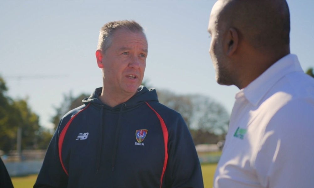 Two men converse on a sunny sports field; one wears a dark blue athletic jacket with a SACA logo, the other in a white polo. Two men converse on a sunny sports field; one wears a dark blue athletic jacket with a SACA logo, the other in a white polo.