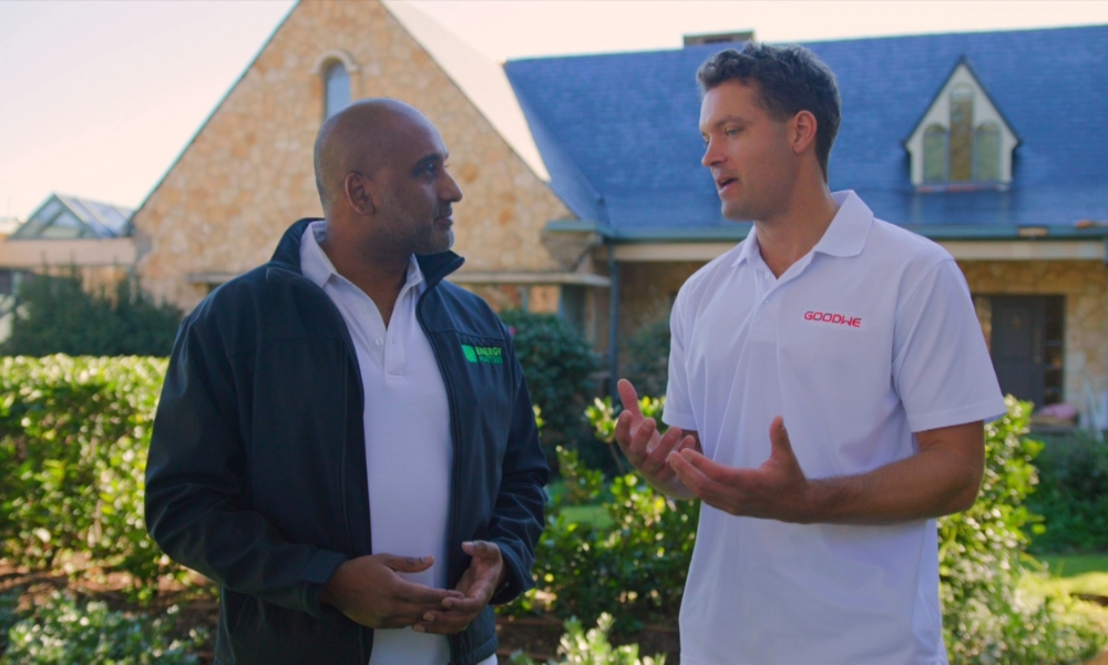 Two men stand outside a stone house, engaged in a conversation, one in a white polo with a logo and the other in a dark jacket. Two men stand outside a stone house, engaged in a conversation, one in a white polo with a logo and the other in a dark jacket.