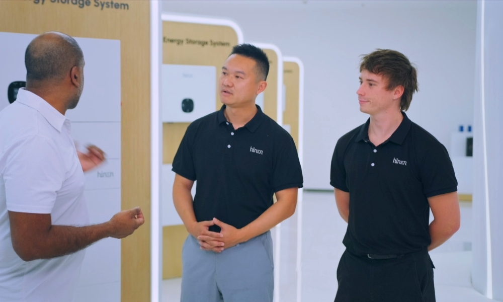 Three men standing and talking in a modern showroom with energy storage units on display behind them, including wood panels labeled 'Energy Storage System'. Three men standing and talking in a modern showroom with energy storage units on display behind them, including wood panels labeled 'Energy Storage System'.