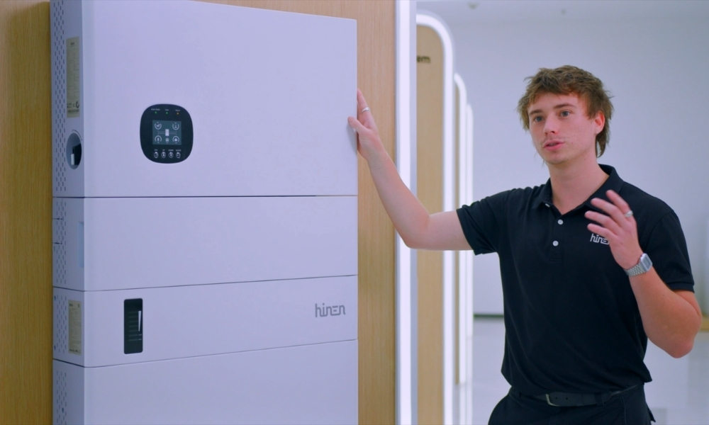 Man in a black polo explains a large white cabinet-like machine, touching its side in a hallway. Man in a black polo explains a large white cabinet-like machine, touching its side in a hallway.