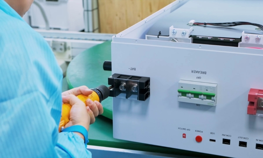 Person in a blue sleeve plugs a yellow-handled connector into a gray electrical control panel on a green worktable. Person in a blue sleeve plugs a yellow-handled connector into a gray electrical control panel on a green worktable.