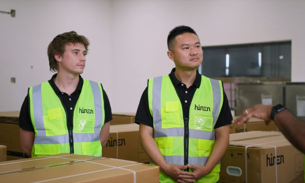 Two men in neon safety vests stand in a warehouse among cardboard boxes, listening to someone off-camera. Two men in neon safety vests stand in a warehouse among cardboard boxes, listening to someone off-camera.