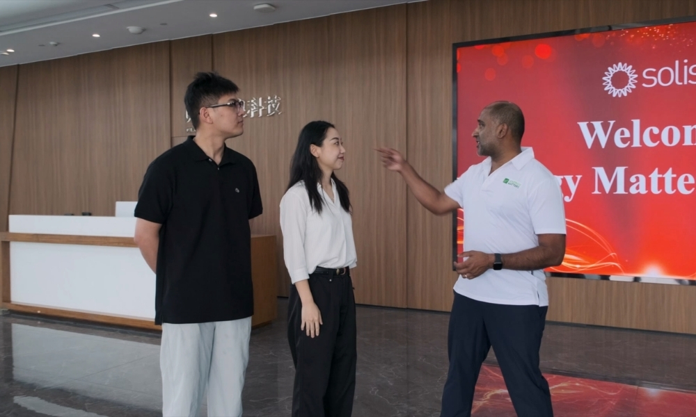 Three people stand in a modern lobby as a man in a white polo explains something to a woman and a man in black shirt, near a red welcome screen. Three people stand in a modern lobby as a man in a white polo explains something to a woman and a man in black shirt, near a red welcome screen.