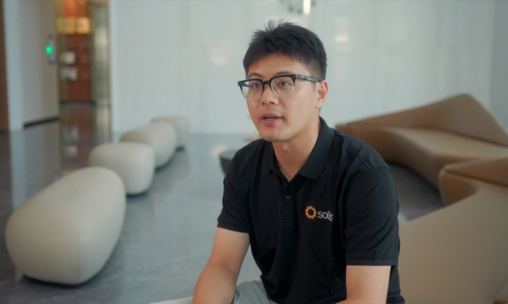 Man wearing a black polo with a Solis logo sits in a bright, minimalist lobby on a curved beige sofa, speaking softly. Man wearing a black polo with a Solis logo sits in a bright, minimalist lobby on a curved beige sofa, speaking softly.