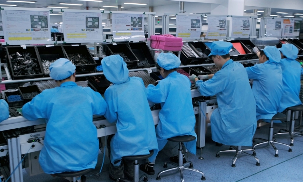 Workers in blue lab coats and caps sit on stools along an electronics assembly line, sorting and assembling components from labeled bins. Workers in blue lab coats and caps sit on stools along an electronics assembly line, sorting and assembling components from labeled bins.
