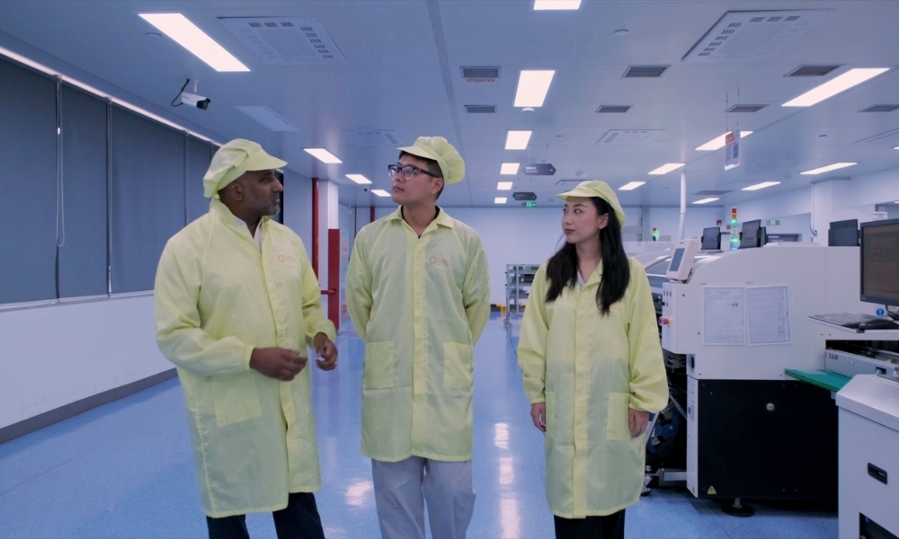Three professionals in yellow cleanroom suits and caps stand in a bright, modern lab, discussing near manufacturing equipment. Three professionals in yellow cleanroom suits and caps stand in a bright, modern lab, discussing near manufacturing equipment.