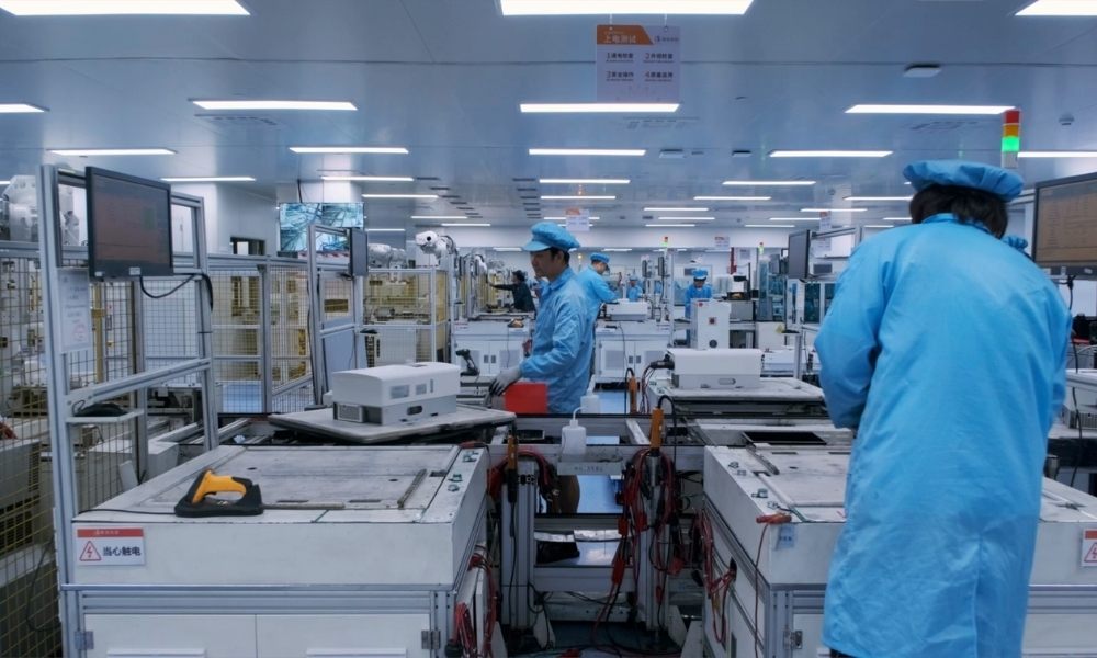 Workers in blue cleanroom suits and caps operate electronics manufacturing equipment in a bright, orderly factory. Workers in blue cleanroom suits and caps operate electronics manufacturing equipment in a bright, orderly factory.
