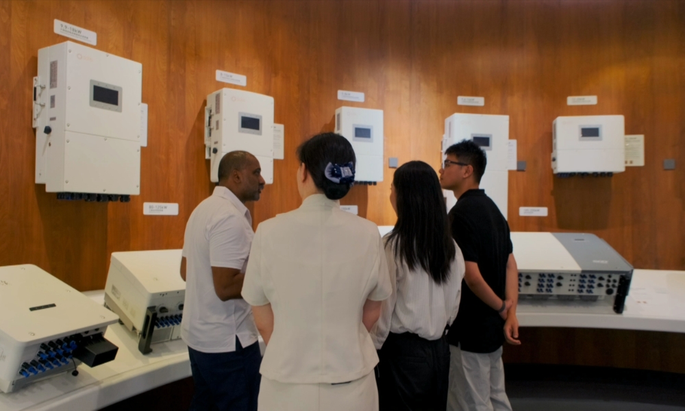 Four people stand in a room, viewing white wall-mounted industrial devices on a wood-paneled backdrop. Four people stand in a room, viewing white wall-mounted industrial devices on a wood-paneled backdrop.